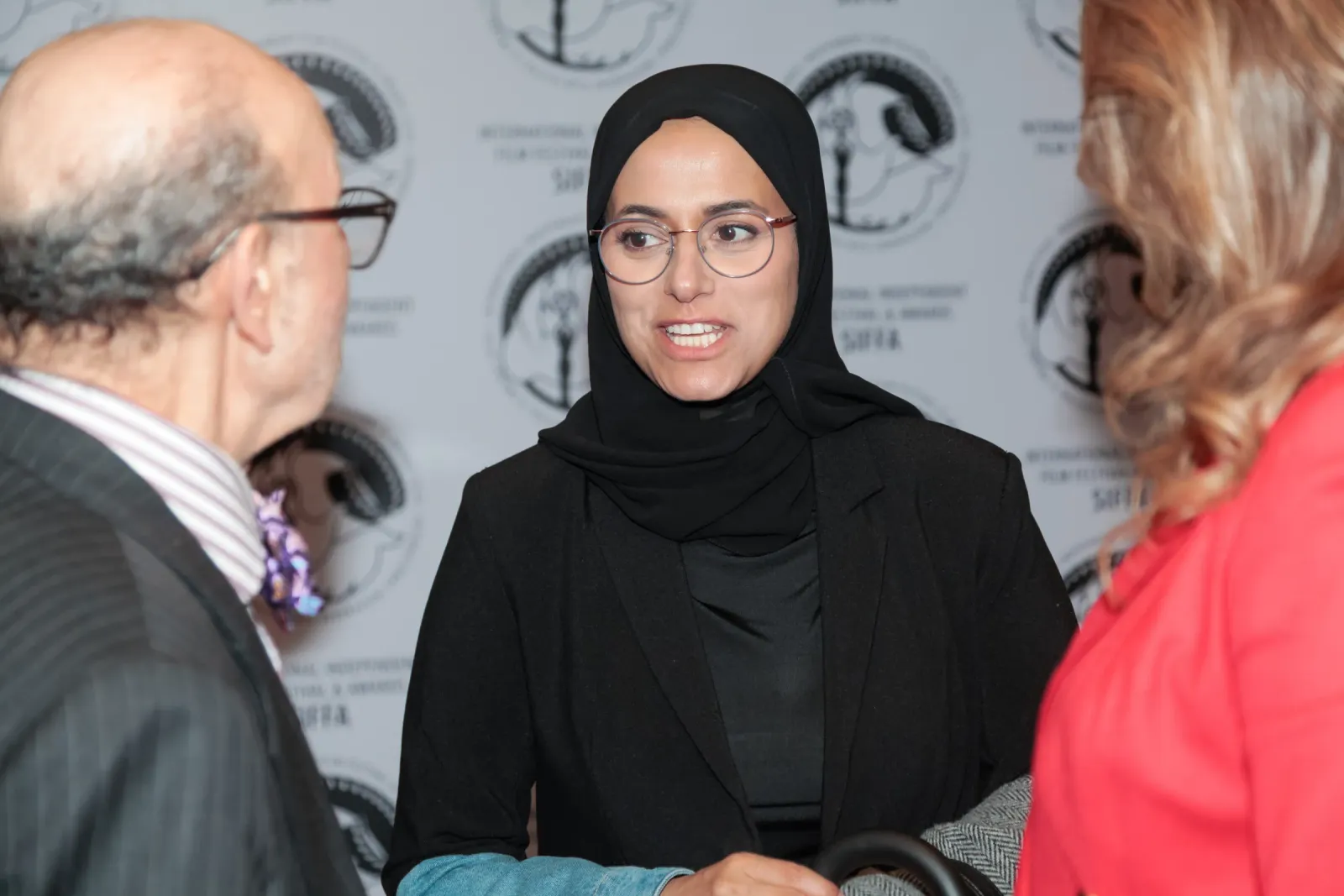 A woman in a black hijab and glasses in conversation with two guests in front of the SIFFA-branded backdrop at the 2024 festival