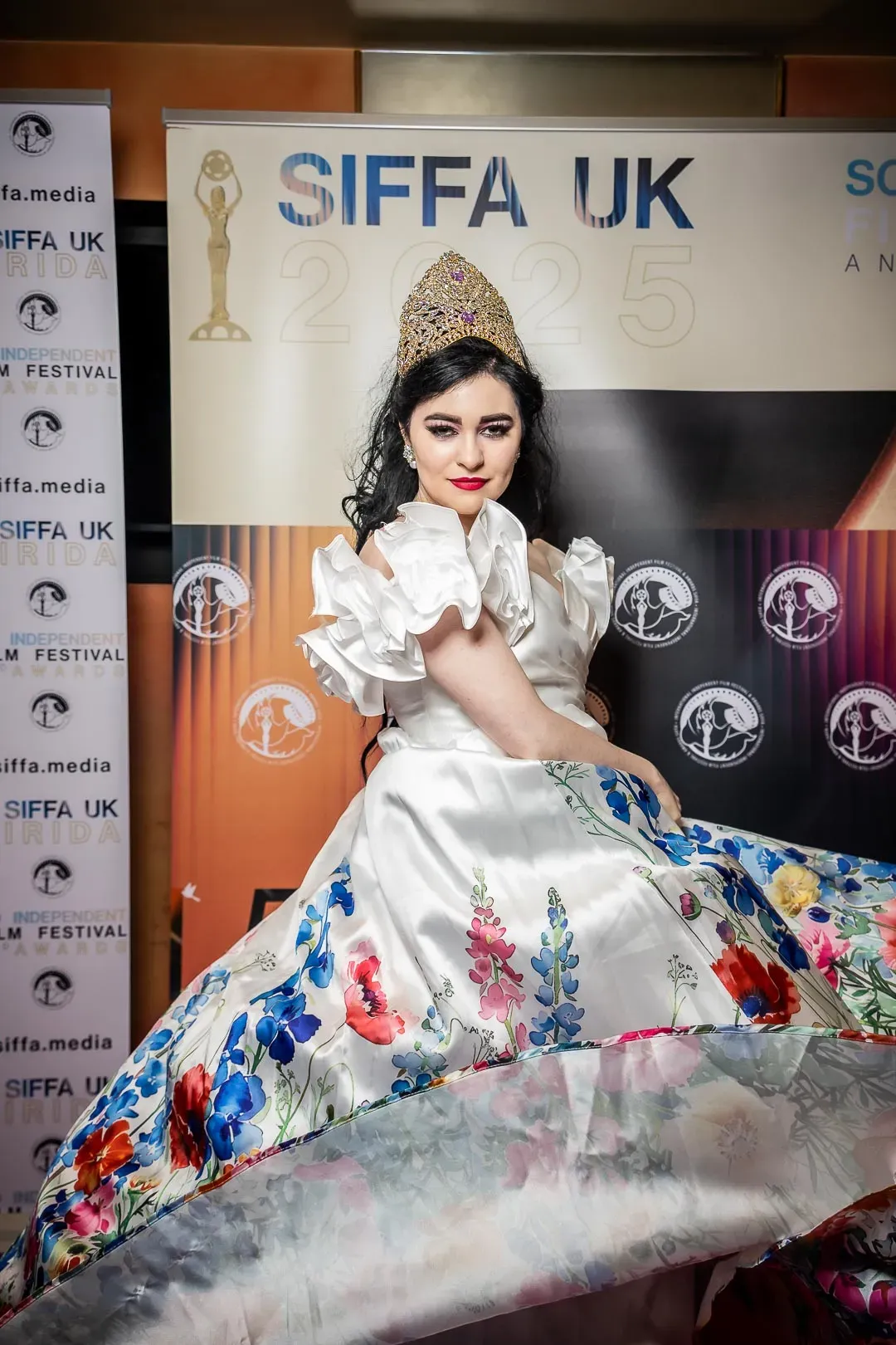 A guest in a jewelled crown and a white gown with a hand-painted floral skirt poses before the SIFFA UK 2025 backdrop