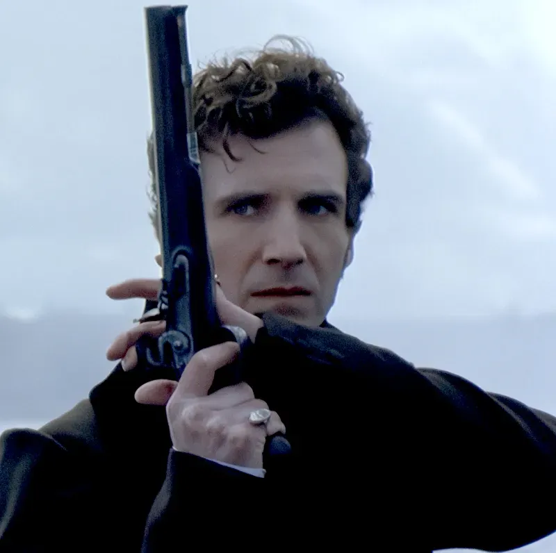Film still: a man in a dark coat raising an antique duelling pistol beside his face against a pale sky