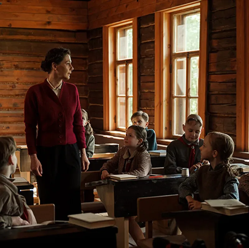 Film still: a teacher in a red cardigan standing before schoolchildren at wooden desks in a rustic log-cabin classroom
